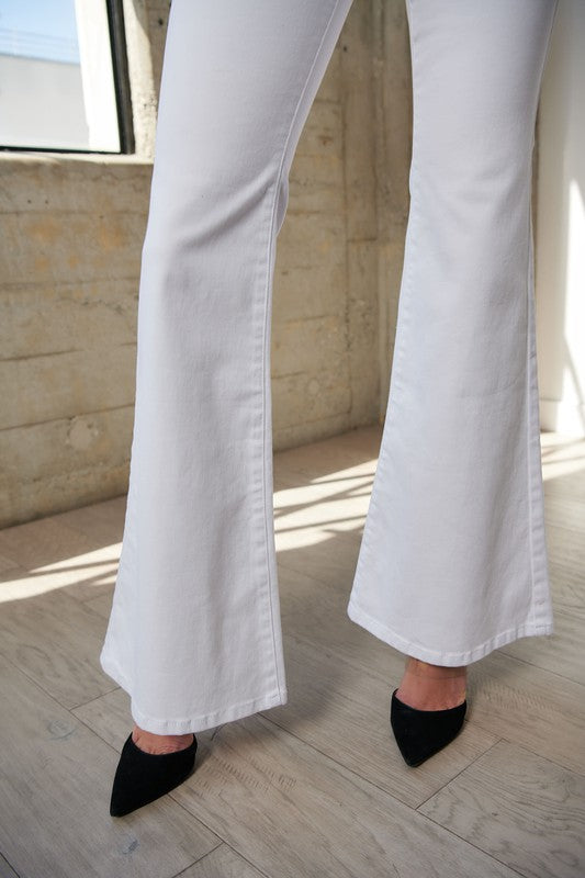 Person wearing high-waisted full-length flare jeans in white, featuring stretch technology, and a black long-sleeve top, standing in a brightly lit room.