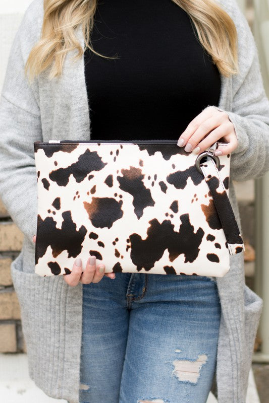 A person flaunts the trending Cow Print Oversized Everyday Clutch, perfectly paired with a cozy oversized gray cardigan, complemented by a black top and ripped jeans.