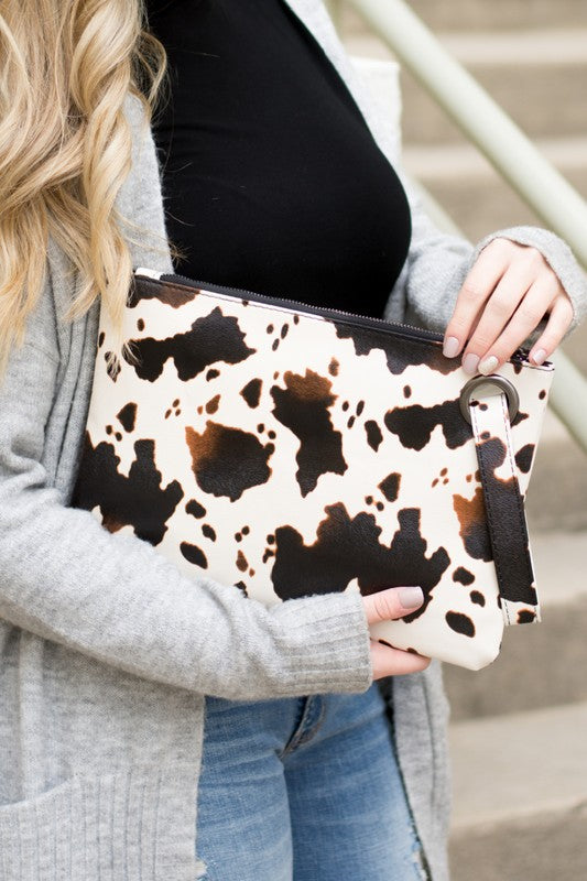 A person flaunts the trending Cow Print Oversized Everyday Clutch, perfectly paired with a cozy oversized gray cardigan, complemented by a black top and ripped jeans.