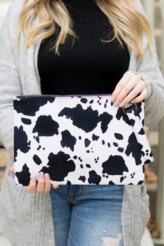 A person flaunts the trending Cow Print Oversized Everyday Clutch, perfectly paired with a cozy oversized gray cardigan, complemented by a black top and ripped jeans.