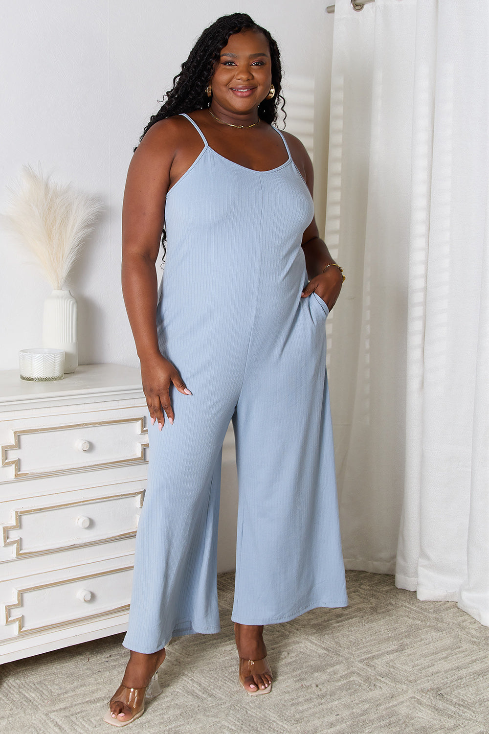 A woman in a stylish Basic Bae Full Size Spaghetti Strap V-Neck Jumpsuit stands indoors near a white dresser adorned with decorative items, smiling with one hand in her pocket.