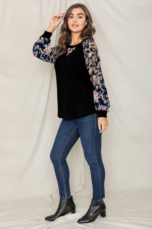 Woman wearing a V-Stitch Floral Raglan Sleeve top, looking at the camera with a neutral expression.