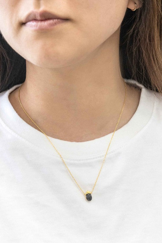 The Pineapple Stone Necklace elegantly showcases a small black oval gemstone pendant on a gold chain, adding a touch of casual jewelry sophistication against the light background.