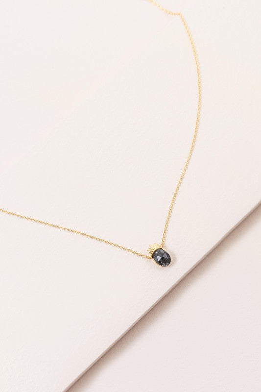 The Pineapple Stone Necklace elegantly showcases a small black oval gemstone pendant on a gold chain, adding a touch of casual jewelry sophistication against the light background.