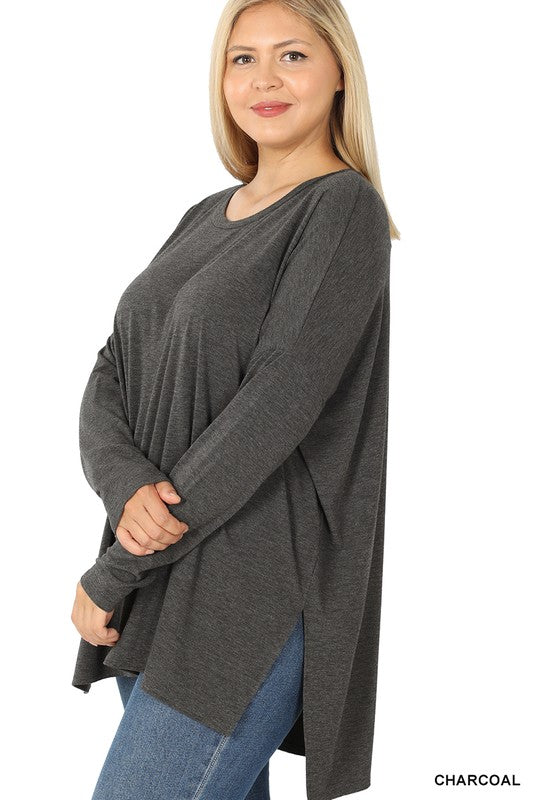 Woman in dark olive Plus Dolman Long Sleeve Round Neck Top and blue jeans standing against a plain background, holding the hem of her top, looking at the camera.