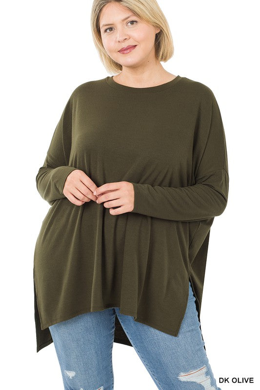 Woman in dark olive Plus Dolman Long Sleeve Round Neck Top and blue jeans standing against a plain background, holding the hem of her top, looking at the camera.