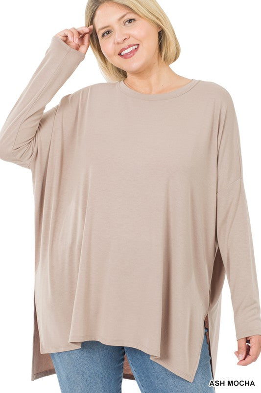Woman in dark olive Plus Dolman Long Sleeve Round Neck Top and blue jeans standing against a plain background, holding the hem of her top, looking at the camera.