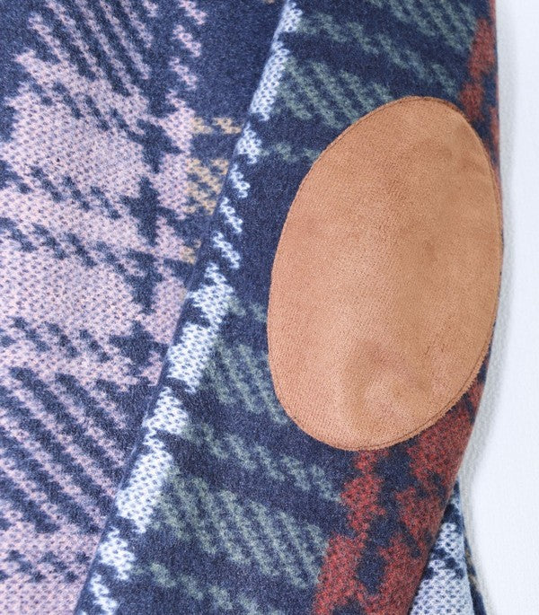 Person with curly hair wearing the Plus Multi Color Plaid Elbow Patch Cardigan over a white shirt and black pants, standing in front of a white paneled wall.