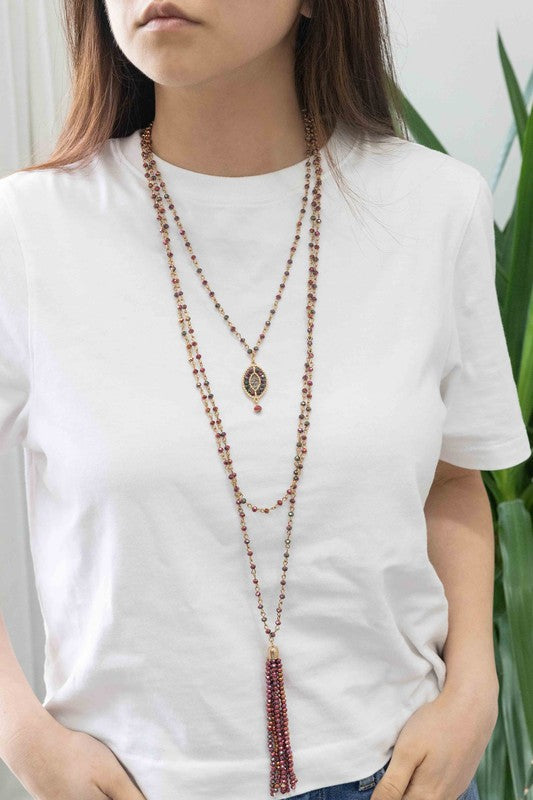 The Angelia Layered Bead Necklace, featuring multiple layers of brass with red beads, pink and green accents, and a decorative pendant on a white background, adds a touch of boho jewelry charm.