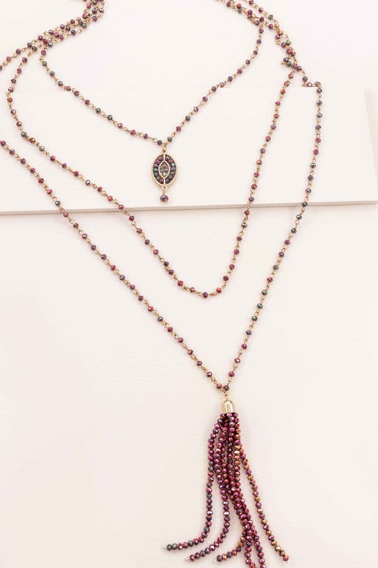 The Angelia Layered Bead Necklace, featuring multiple layers of brass with red beads, pink and green accents, and a decorative pendant on a white background, adds a touch of boho jewelry charm.