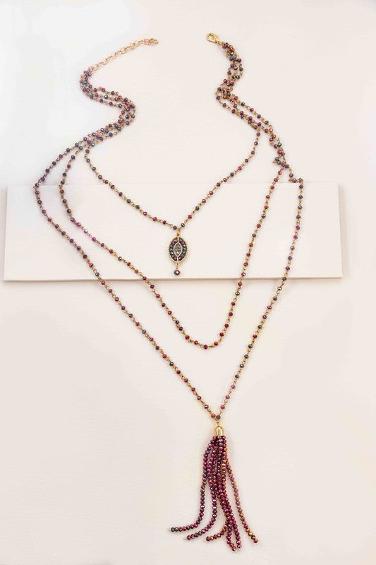 The Angelia Layered Bead Necklace, featuring multiple layers of brass with red beads, pink and green accents, and a decorative pendant on a white background, adds a touch of boho jewelry charm.