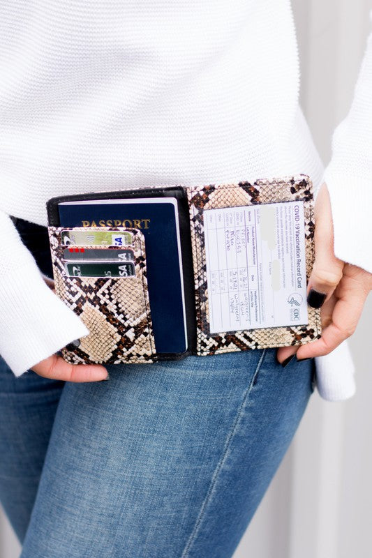 A person wearing a white sweater and blue jeans is holding an open Passport and Vaccine Credit Card Wallet in leopard print, showcasing a passport, vaccine card holder, cards, and a boarding pass.