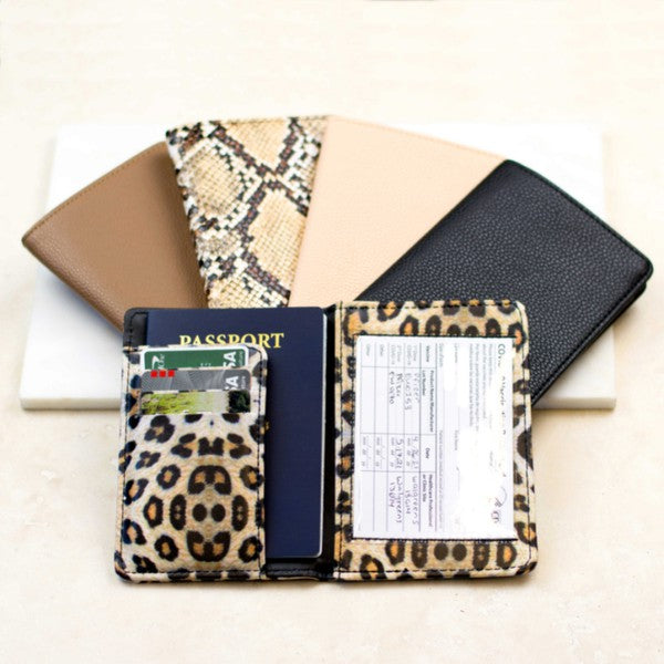 A person wearing a white sweater and blue jeans is holding an open Passport and Vaccine Credit Card Wallet in leopard print, showcasing a passport, vaccine card holder, cards, and a boarding pass.