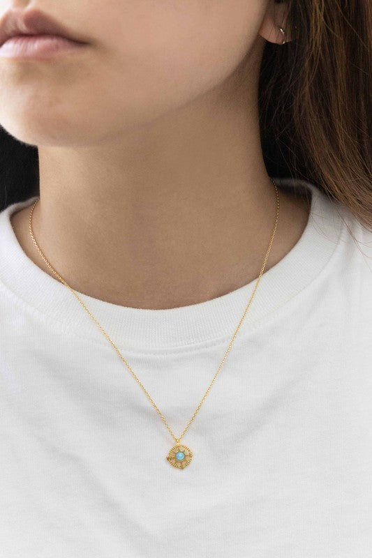 The Pixum Compass Stone Necklace, featuring a thin gold chain and a pendant with a turquoise stone set in a circular gold disc, captures the bohemian jewelry essence beautifully against a light background.