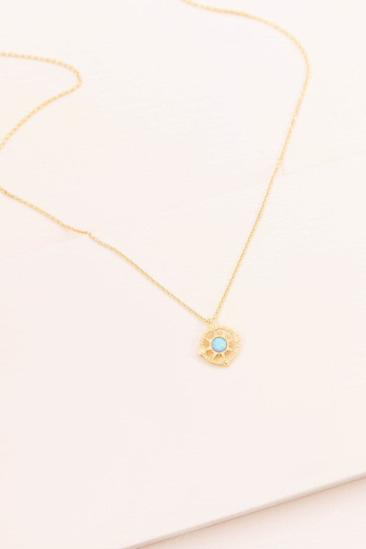 The Pixum Compass Stone Necklace, featuring a thin gold chain and a pendant with a turquoise stone set in a circular gold disc, captures the bohemian jewelry essence beautifully against a light background.