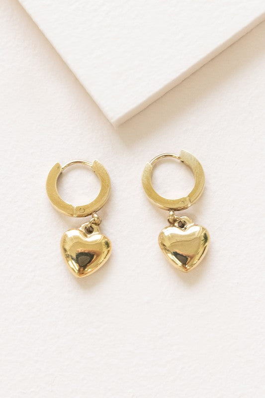 The Adore Hoop Earrings are crafted from stainless steel, plated in 14k gold, and feature charming heart-shaped pendants that dangle elegantly against a pristine white background.