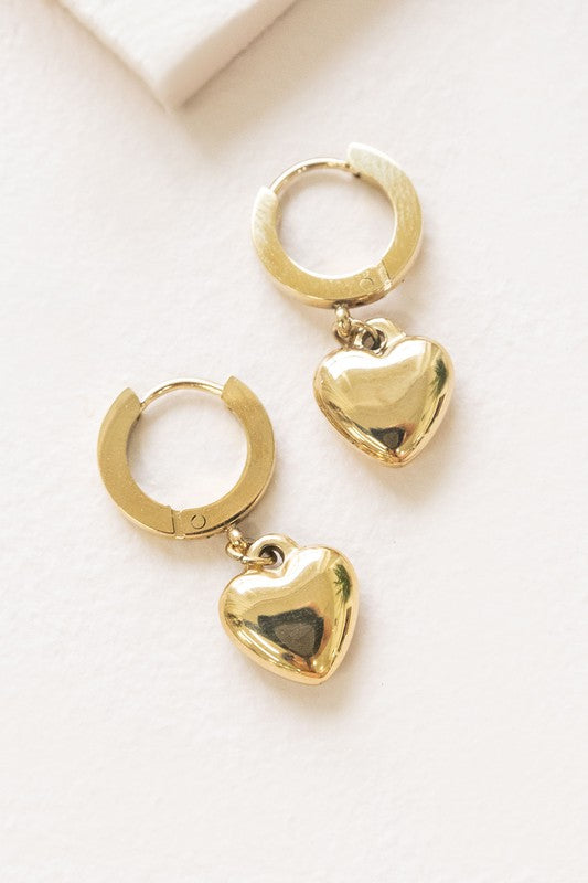 The Adore Hoop Earrings are crafted from stainless steel, plated in 14k gold, and feature charming heart-shaped pendants that dangle elegantly against a pristine white background.