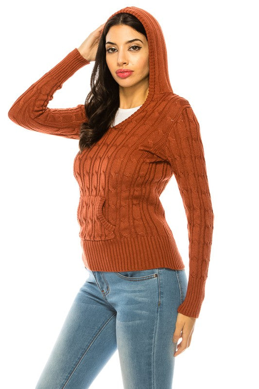 A woman stands wearing a rust-colored knit hoodie sweater and blue jeans. She has long dark hair and is facing the camera with a neutral expression.