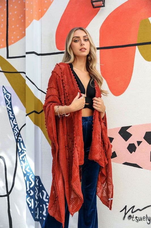 A woman stands in front of a colorful mural. She wears an Embroidered Zig Zag Soft Kimono with pom-poms, a black top, and blue pants. She has long blonde hair and is looking directly at the camera.