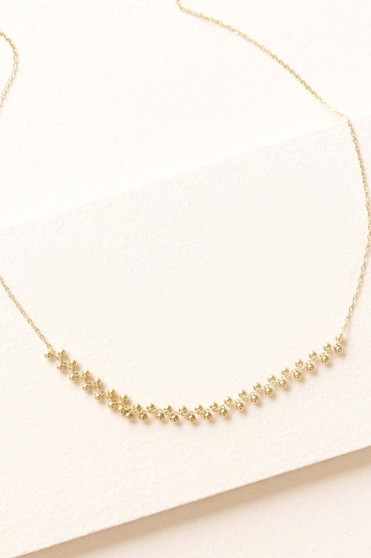 The Samba Charm Necklace is an elegant accessory adorned with small, evenly spaced floral-shaped charms that rest gracefully on a white background. Made from 14k gold-plated stainless steel, this necklace embodies timeless style and sophistication.