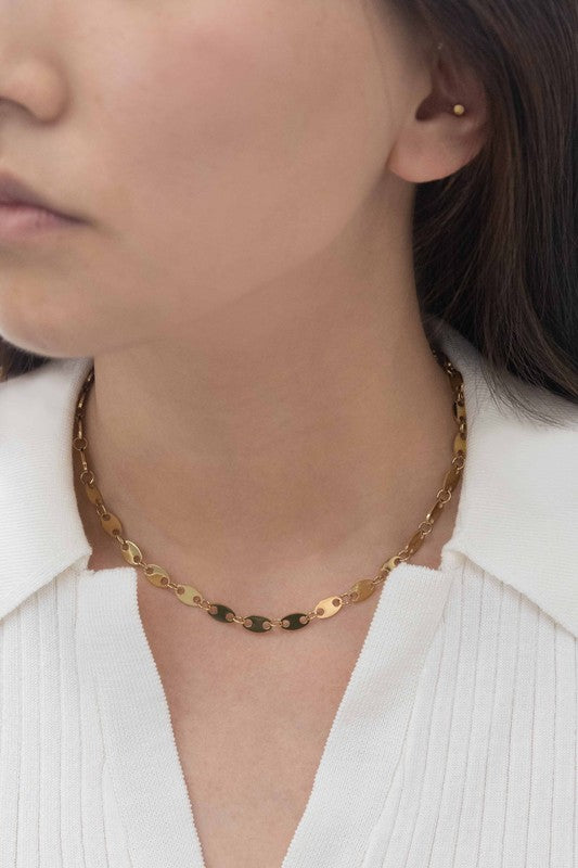 The Oval Chain Necklace, featuring a 14k gold plated finish with oval links and an adjustable clasp, is elegantly displayed on a white surface, highlighting its sophisticated design and stainless steel durability.