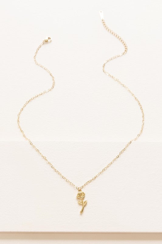 The La Vie En Rose Charm Necklace boasts a gold pendant intricately designed in the form of a rose, elegantly hanging against a subtle background. This classic accessory is 14k gold plated, providing sophistication to any outfit.