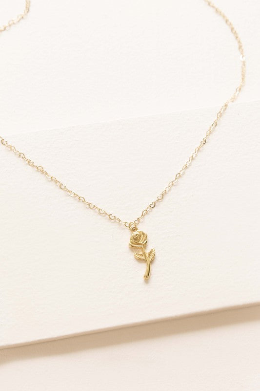 The La Vie En Rose Charm Necklace boasts a gold pendant intricately designed in the form of a rose, elegantly hanging against a subtle background. This classic accessory is 14k gold plated, providing sophistication to any outfit.