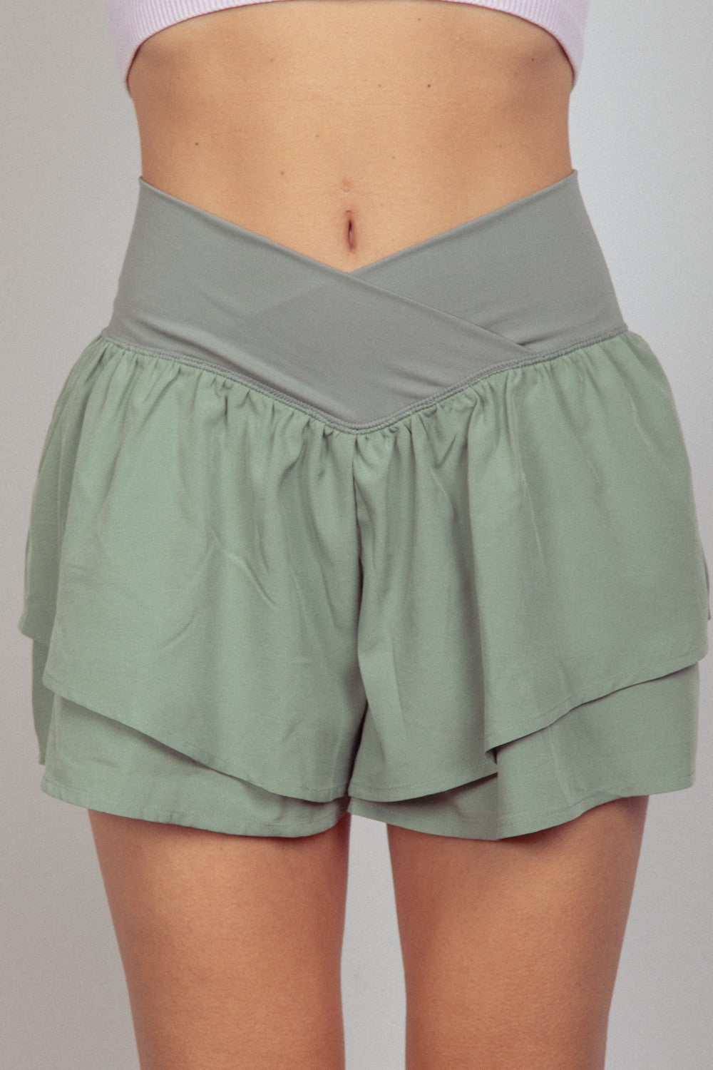 A close-up of a person wearing a light purple cropped top and sage green VERY J V-Shaped High Waist Layered Active Shorts with a crossover waistband.