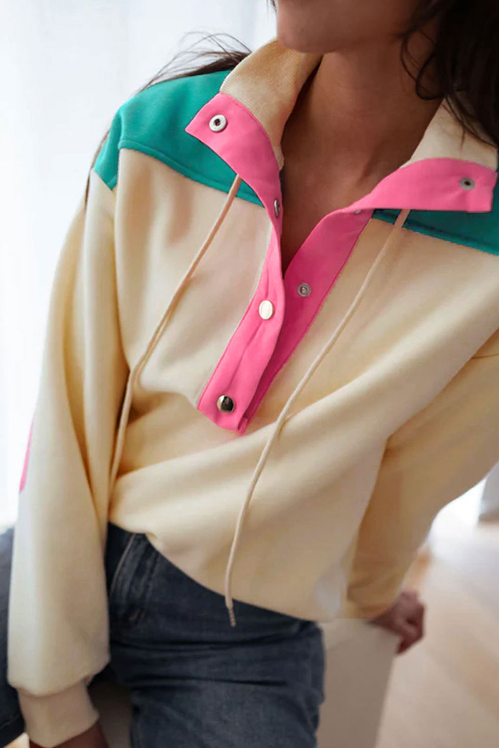 A woman wearing the Apricot Color Block Elbow Patch Half Button Sweatshirt, featuring cream and mint colors with pink elbow patches and a turn-down collar with snap button closure, stands outdoors. She has long wavy hair, sunglasses, a hat, and is wearing blue jeans.