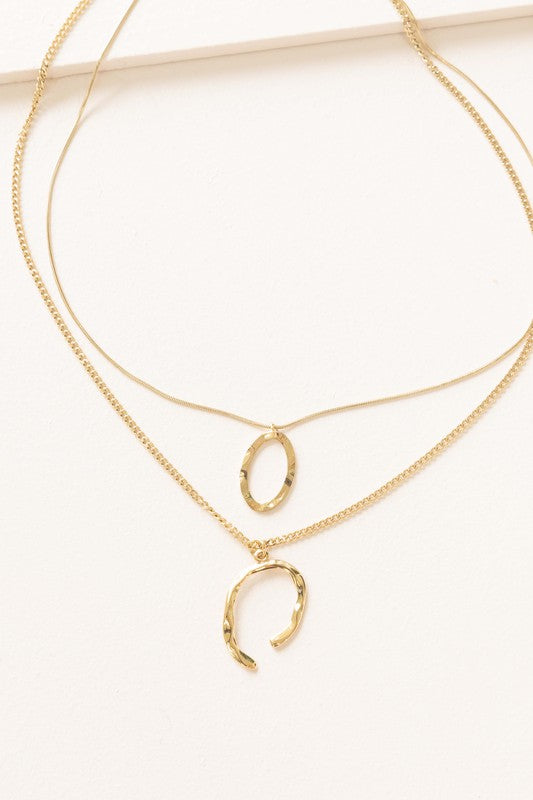 Introducing the Ithaca Layered Necklace, a stunning piece crafted from 14k gold plating. This necklace features two delicate chains adorned with a small oval pendant and a chic horseshoe pendant.