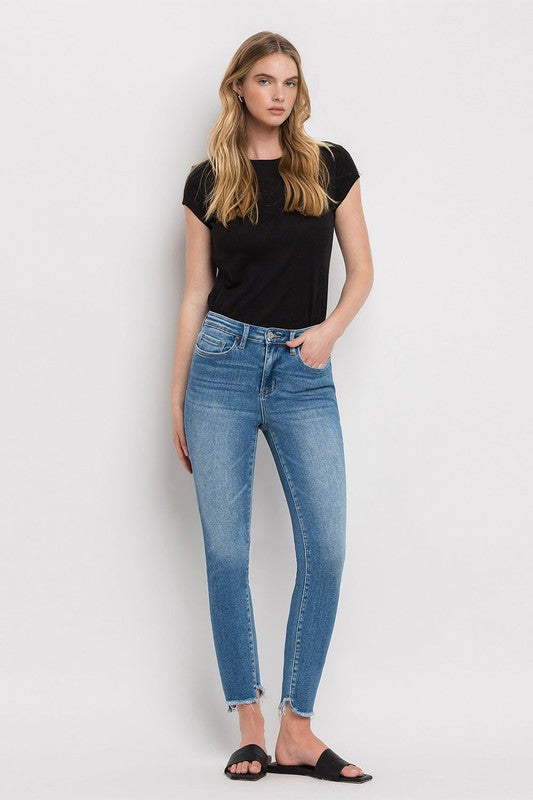 Person wearing blue High Rise Raw Step Hem Ankle Skinny Jeans, a black top, and black slide sandals. One hand is in their pocket.