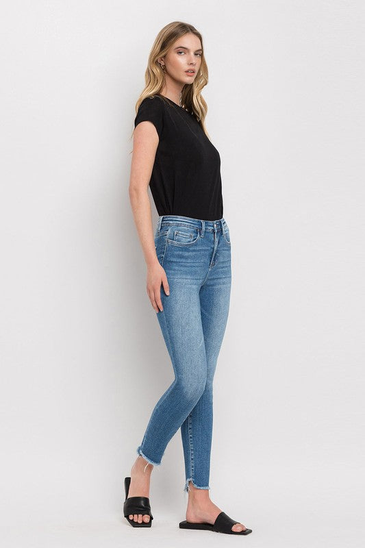 Person wearing blue High Rise Raw Step Hem Ankle Skinny Jeans, a black top, and black slide sandals. One hand is in their pocket.