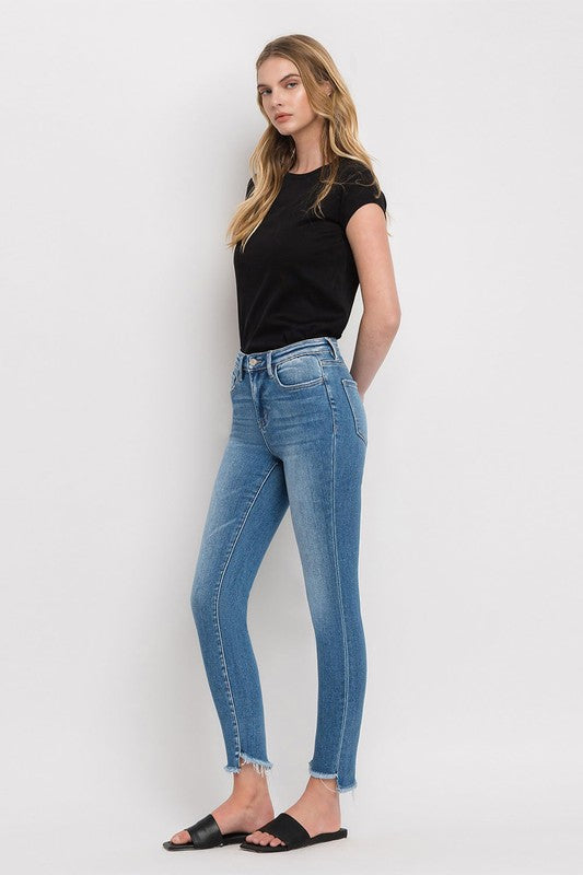 Person wearing blue High Rise Raw Step Hem Ankle Skinny Jeans, a black top, and black slide sandals. One hand is in their pocket.