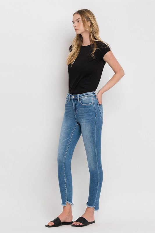 Person wearing blue High Rise Raw Step Hem Ankle Skinny Jeans, a black top, and black slide sandals. One hand is in their pocket.