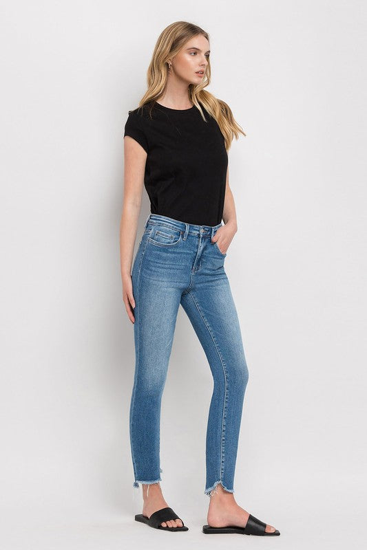 Person wearing blue High Rise Raw Step Hem Ankle Skinny Jeans, a black top, and black slide sandals. One hand is in their pocket.