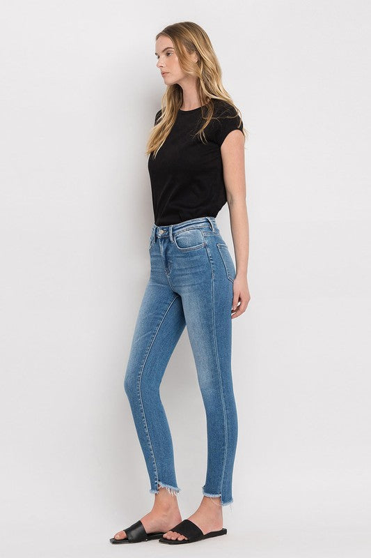 Person wearing blue High Rise Raw Step Hem Ankle Skinny Jeans, a black top, and black slide sandals. One hand is in their pocket.