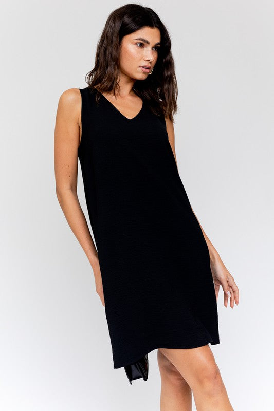 A woman elegantly poses in the Sleeveless V-Neck Dress, highlighting a chic and stylish appearance against a minimalistic backdrop. This versatile wardrobe piece epitomizes both elegance and simplicity.