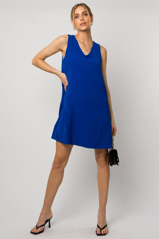 A woman elegantly poses in the Sleeveless V-Neck Dress, highlighting a chic and stylish appearance against a minimalistic backdrop. This versatile wardrobe piece epitomizes both elegance and simplicity.