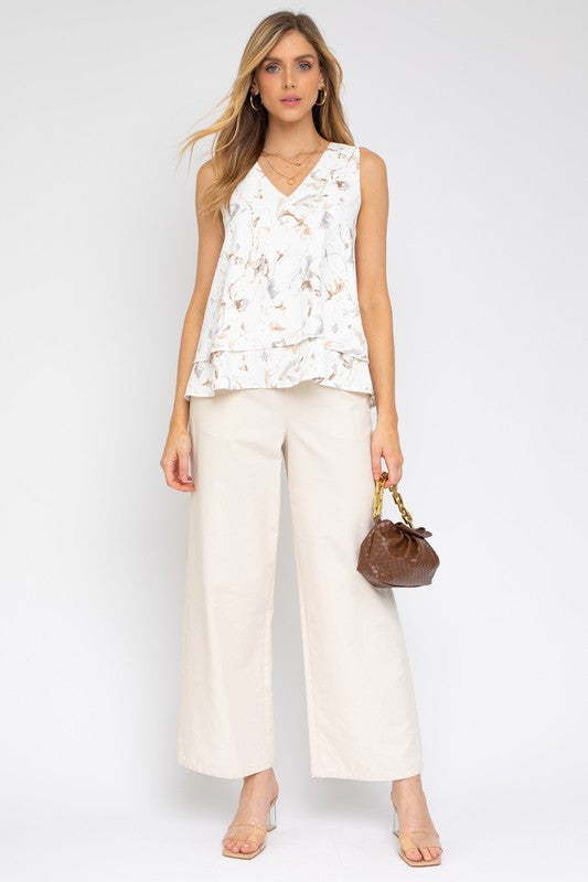 A woman wearing a Sleeveless V-Neck Layered Bottom Abstract Top and cream pants stands against a plain background, holding a small handbag.