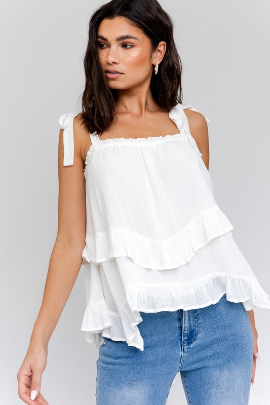 Woman wearing a Sleeveless Asymmetrical Hem Line Tiered Top and blue jeans, standing against a plain background.