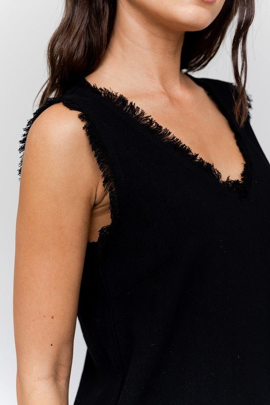 Dressed in a stylish Edgy Frayed Top paired with blue jeans, an individual sits on a stool against a simple backdrop, poised for casual outings.