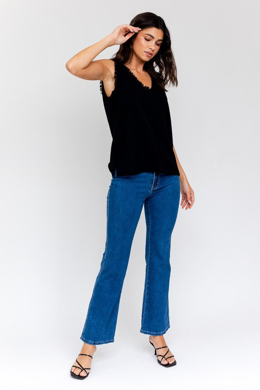 Dressed in a stylish Edgy Frayed Top paired with blue jeans, an individual sits on a stool against a simple backdrop, poised for casual outings.