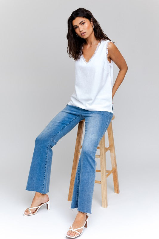 Dressed in a stylish Edgy Frayed Top paired with blue jeans, an individual sits on a stool against a simple backdrop, poised for casual outings.