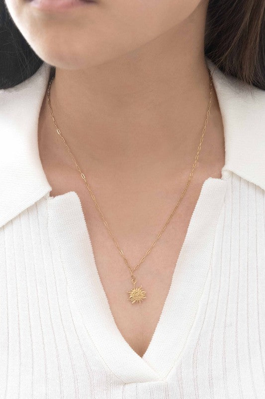 The Helio Charm Necklace is a piece made with 14k gold plating, featuring a sun-shaped pendant with a smiling face against a light backdrop.