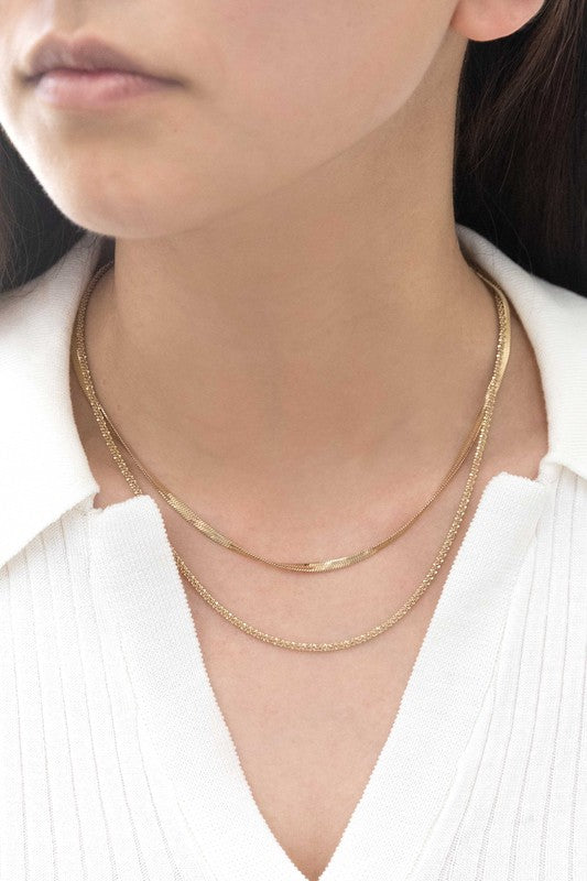 The Laertes Layered Necklace, meticulously crafted with 14k gold plating and durable stainless steel, features a delicate chain paired with a flat herringbone chain. It is elegantly displayed on a light surface.