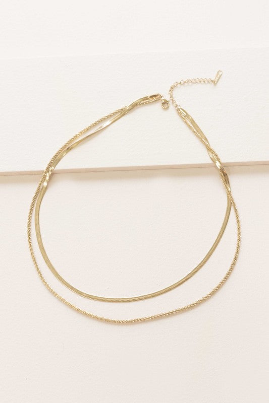 The Laertes Layered Necklace, meticulously crafted with 14k gold plating and durable stainless steel, features a delicate chain paired with a flat herringbone chain. It is elegantly displayed on a light surface.