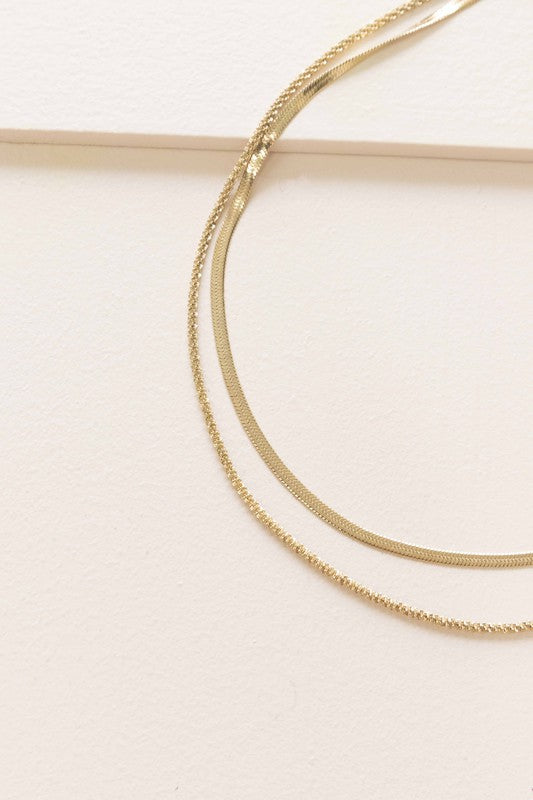 The Laertes Layered Necklace, meticulously crafted with 14k gold plating and durable stainless steel, features a delicate chain paired with a flat herringbone chain. It is elegantly displayed on a light surface.