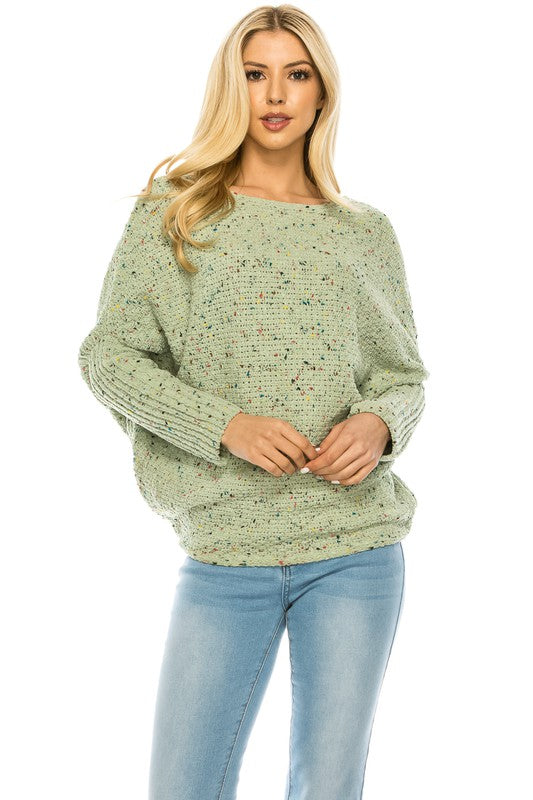 A woman with long blonde hair wears a multi-color speckled knit jumper and light blue jeans. She poses with one hand on her hip and a relaxed expression on her face.