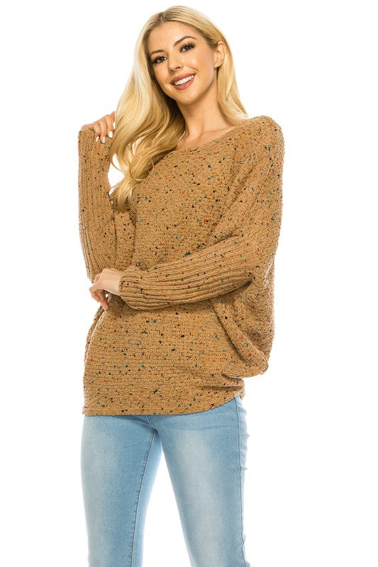 A woman with long blonde hair wears a multi-color speckled knit jumper and light blue jeans. She poses with one hand on her hip and a relaxed expression on her face.