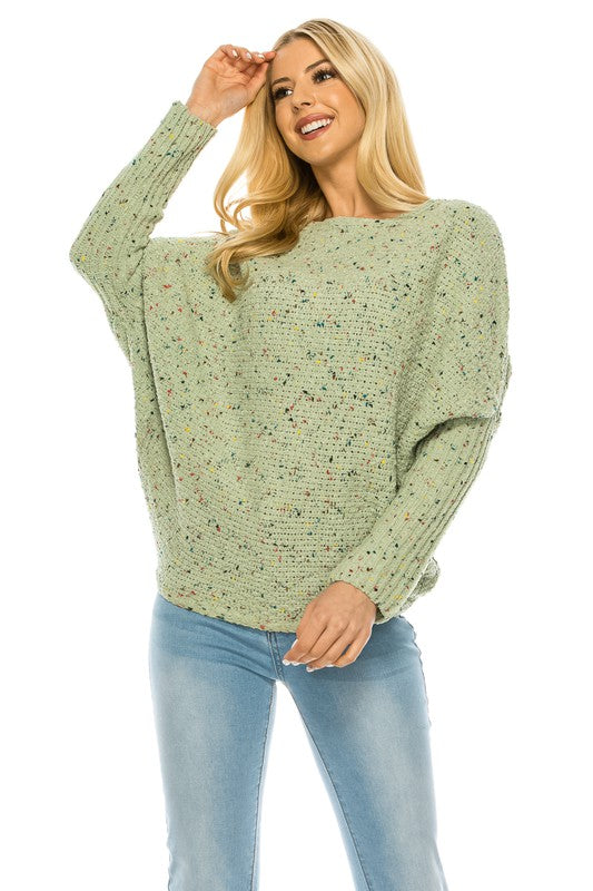 A woman with long blonde hair wears a multi-color speckled knit jumper and light blue jeans. She poses with one hand on her hip and a relaxed expression on her face.